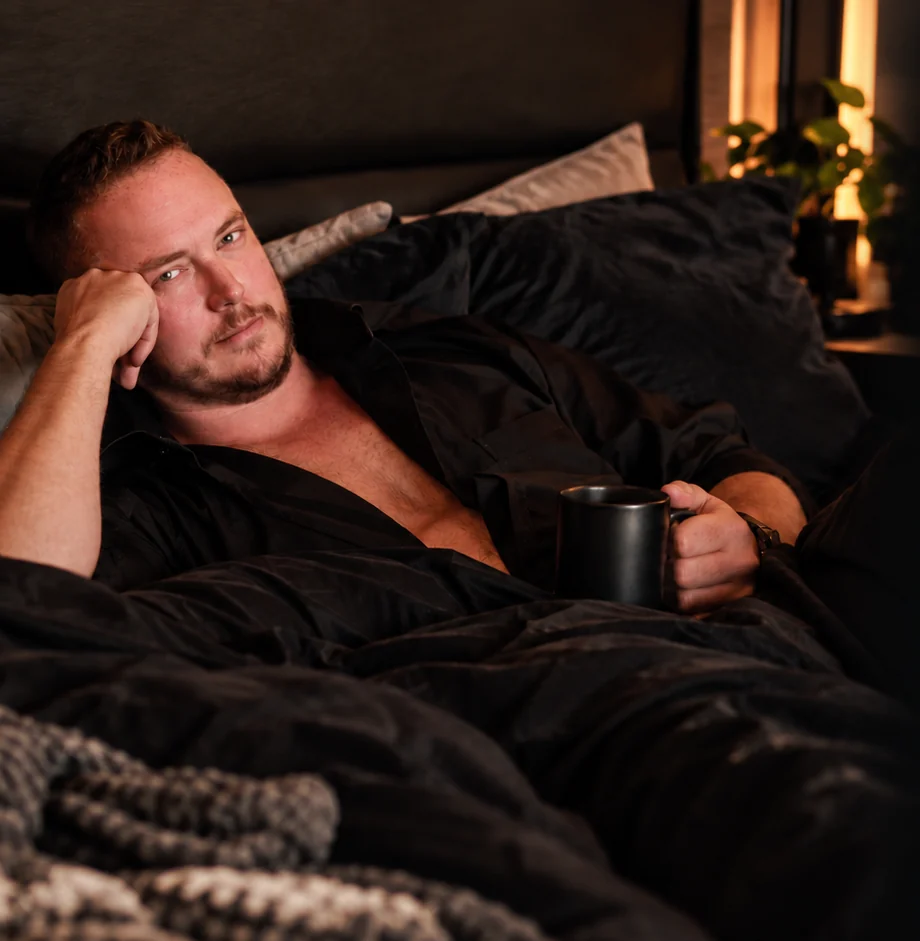 Ayce Kyptyn resting in a dark bed with a mug in hand, looking toward the camera