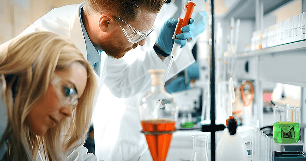 Scientists in a lab, wearing goggles and gloves. One is using a pipette with red liquid in a flask. Background has lab equipment.