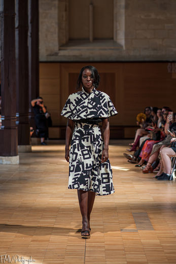 Mannequin présentant une robe à imprimé graphique noir et blanc, accompagnée d'une cape assortie, lors d'un défilé de mode.