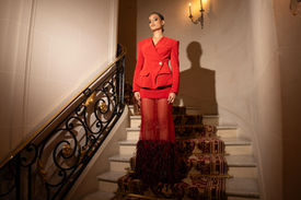 Mannequin photographiée en contre-plongée au milieu d'un escalier d'honneur, vêtue d'un ensemble de créateur rouge vif structuré et d'une jupe transparente à plumes.