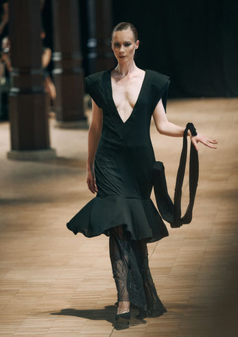 Une mannequin défile sur le podium dans une robe de soirée de luxe noire à col en V, avec un ourlet évasé et un châle assorti dans le même tissu.