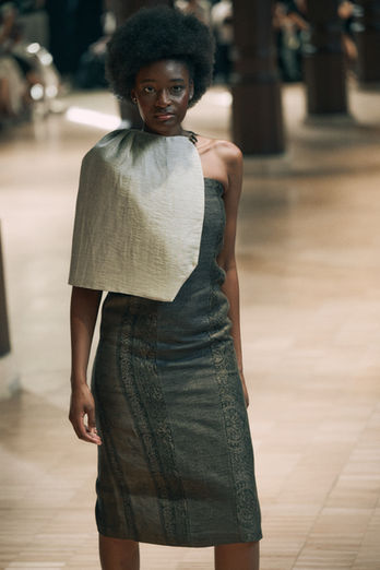 Mannequin avec une coupe afro portant une robe de créateur grise rehaussée d'un drapé volumineux asymétrique sur l'épaule.