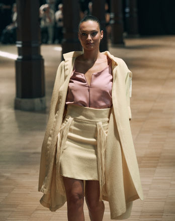 Mannequin sur un podium portant un long manteau jaune ouvert sur un top rose pastel et une jupe assortie à franges.