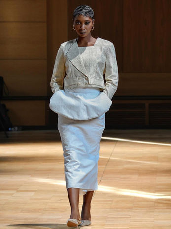 Un mannequin défile sur un podium, vêtue d'une veste courte structurée de couleur blanc cassé et d'une jupe longue.