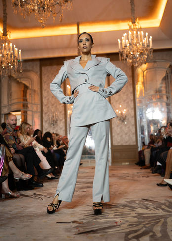 Mannequin posant les mains sur les hanches dans un ensemble tailleur-pantalon d'un gris bleuté très clair, structuré par un haut asymétrique à boutons.