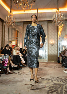 Mannequin défilant dans un ensemble jacquard texturé aux nuances sombres et reflets métalliques bleutés, composé d'une veste courte et d'une jupe crayon.