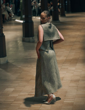 Vue arrière d'un mannequin posant sur un podium en longue robe texturée vert olive dotée d'un grand col géométrique structuré.