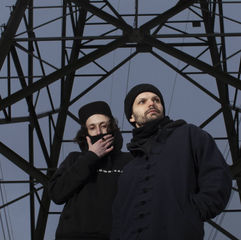 Music producers alllone stand in black jackets under an electricity pylon.