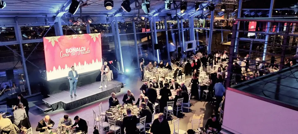 Sala elegante allestita per la cena aziendale Bonaldi, con tavoli apparecchiati e spettacolo live su palco davanti a un maxischermo rosso.