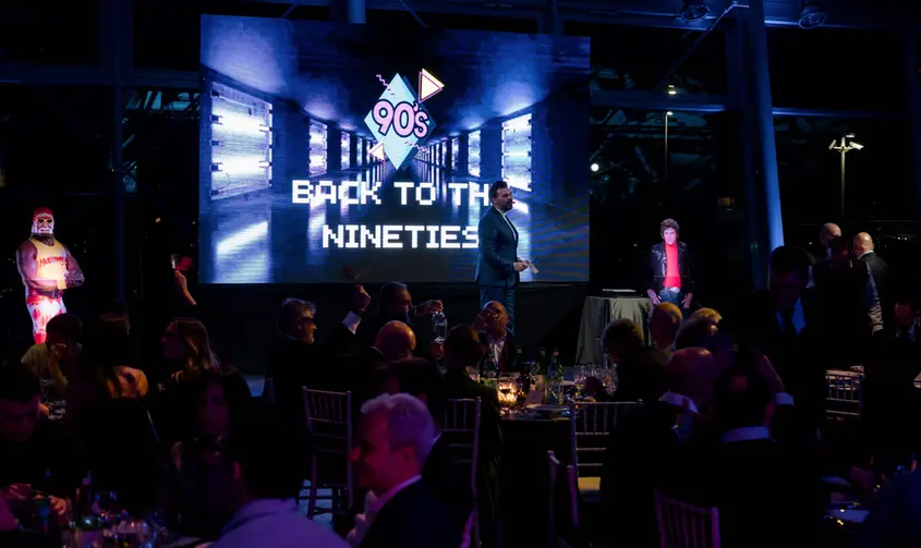 Palco dell’evento “Back to the 90s” con sagome di Hulk Hogan e Michael Knight accanto al conduttore.
