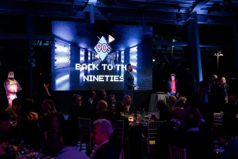 Palco dell’evento “Back to the 90s” con sagome di Hulk Hogan e Michael Knight accanto al conduttore.