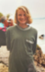 Teenaged Abi stands on a beach smiling and with her hands behind her back. She has chin length wavy blonde hair and is wearing a baggy grey t-shirt.