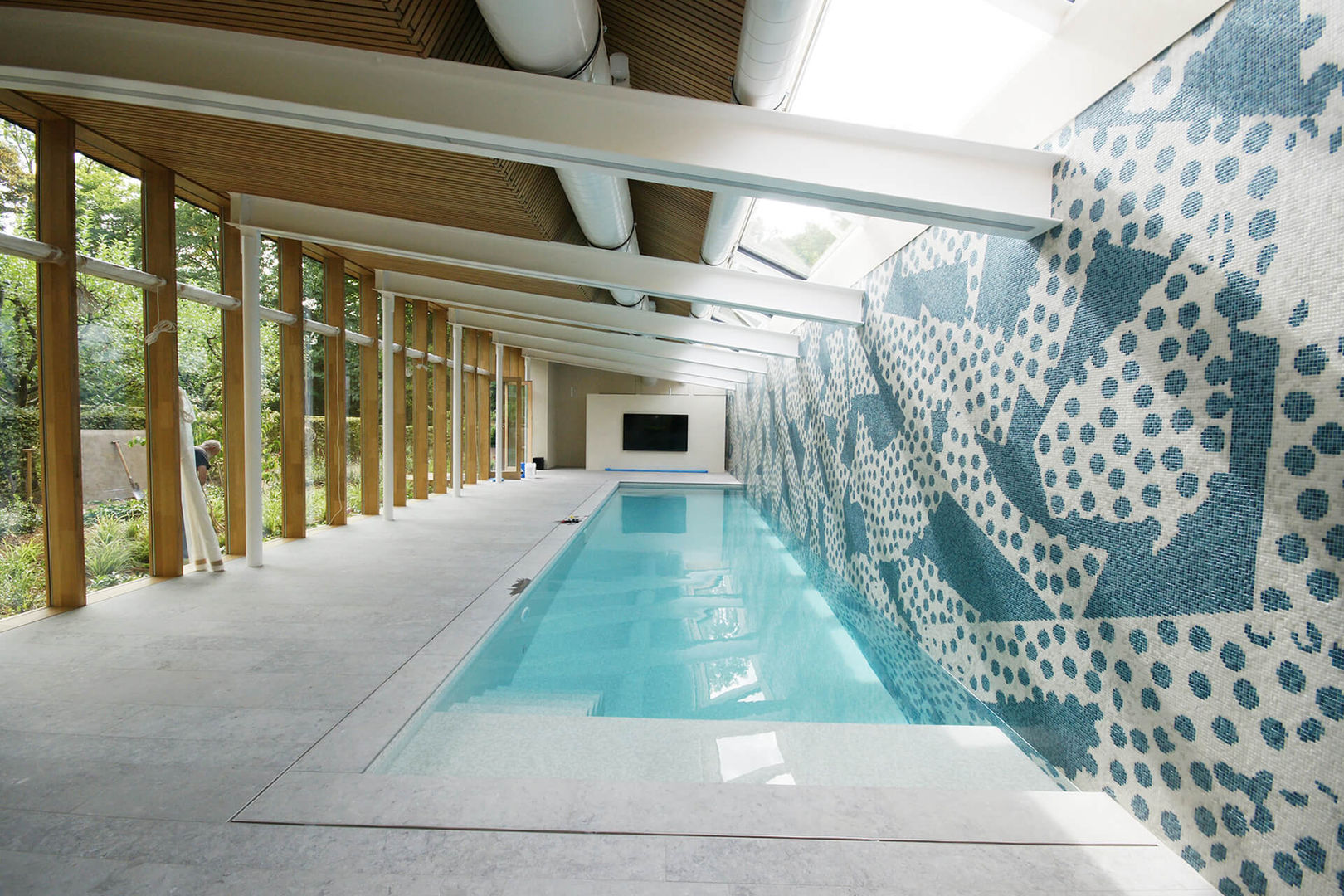 Ce bâtiment a trouvé sa place dans ce beau jardin très naturellement et a permis d’y intégrer la piscine couverte souhaitée par le maître de l’ouvrage.
Il s’agit d’une structure mixte acier et bois qui participe grandement aux qualités spatiales du lieux dont les finitions sont très soignées et affirment les matériaux qui la compose, sol en pierre naturelle, façade vitrée et châssis en chêne ainsi que le plafond acoustique qui este chêne naturel également.
La grande façade vitrée permet un dialogue permanent entre bâtiment et jardin.
Le mur du fond descend dans la piscine qui y est accolée, il est entièrement revêtu d’une fresque en mosaïque réalisée par l’artiste Yves Zurestrassen.
