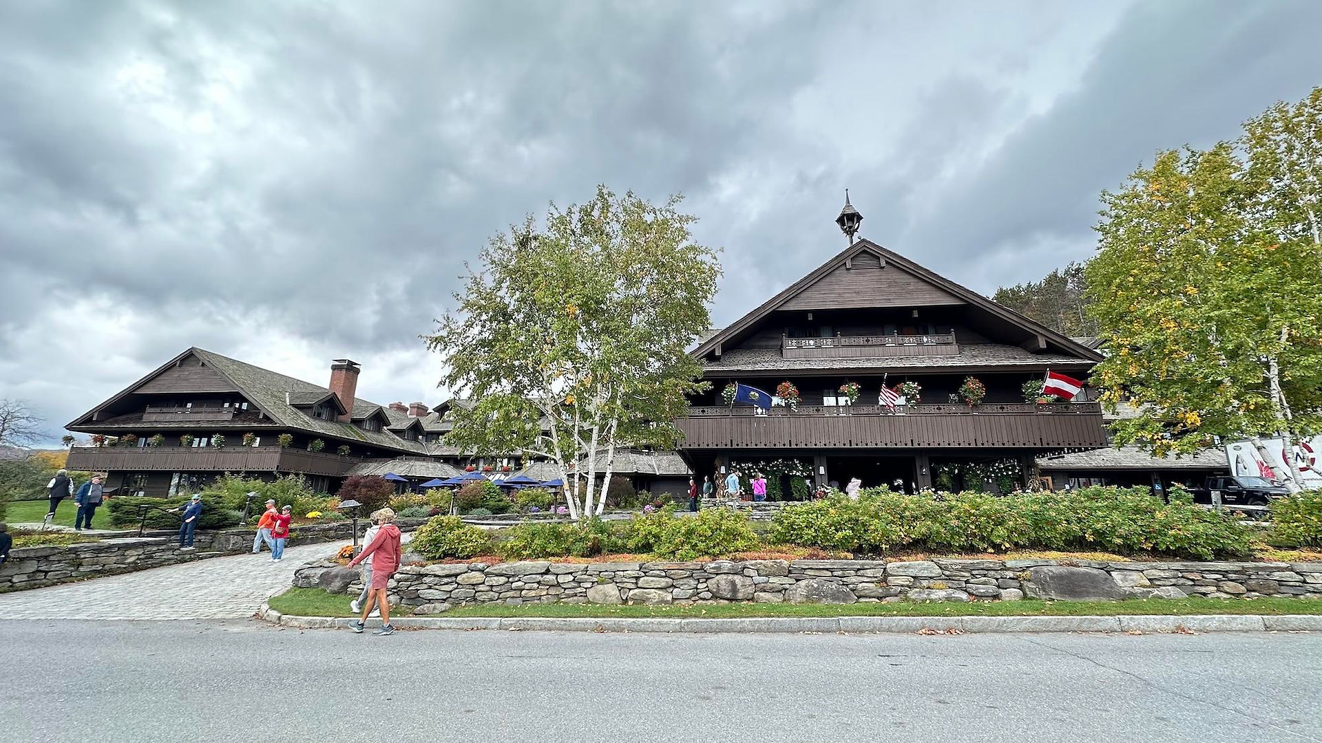 Exterior of the Trapp Family Lodge in Stowe, Vermont, with dark wood balconies, stone landscaping, autumn trees, and Austrian and American flags under a cloudy sky.