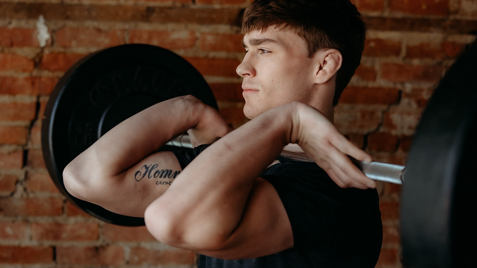 Man lifting a barbell indoors against a brick wall background. Focused expression, tattoo visible on arm.