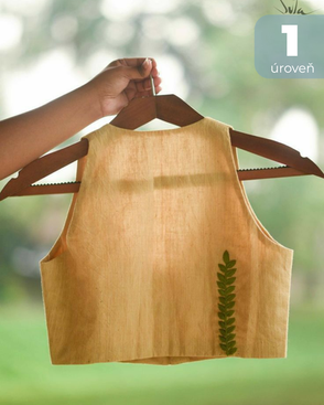 Svetlý detský top bez rukávov s vyšívaným zeleným lístkom zavesený na drevenom vešiaku v prírode.