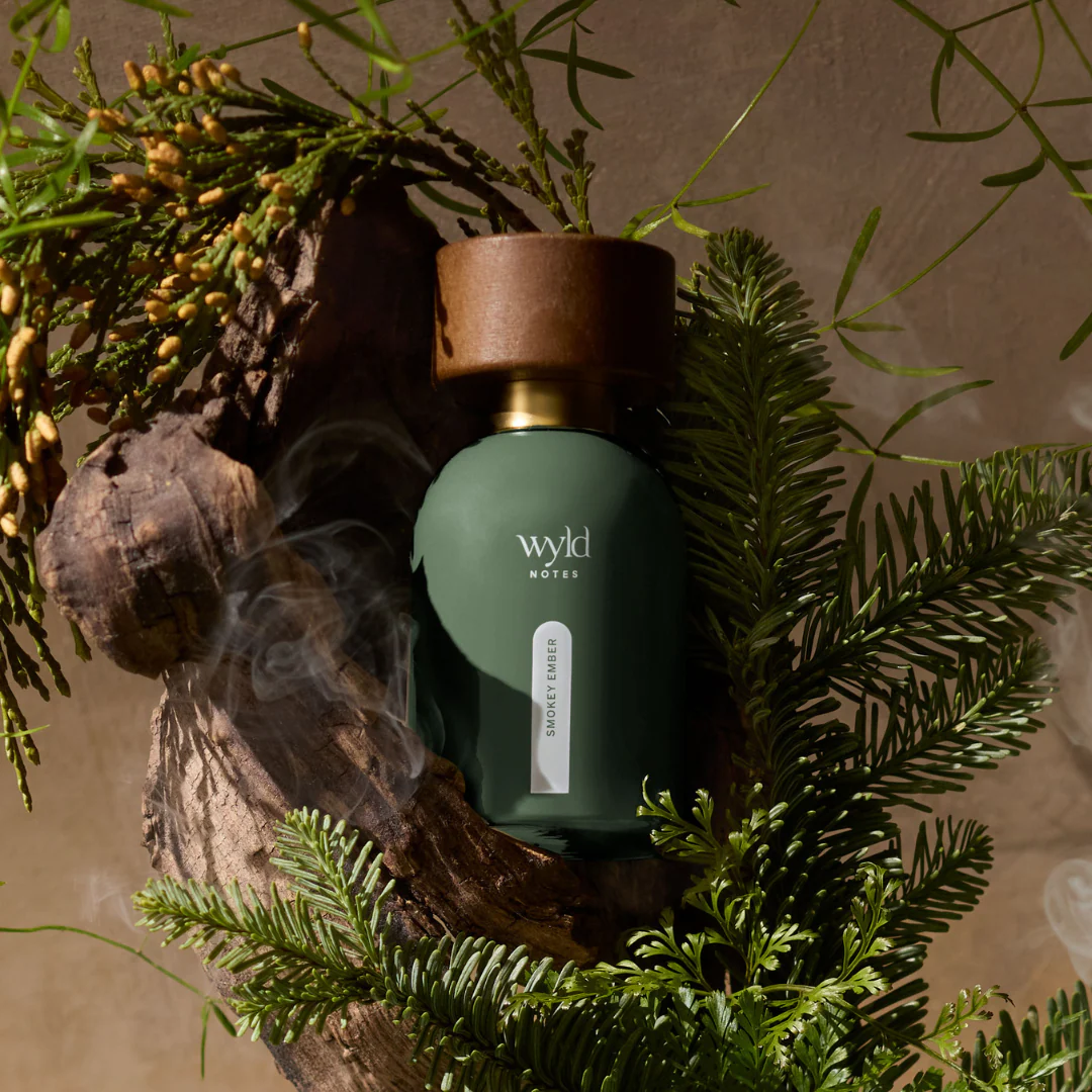 Green perfume bottle labeled "wyld NOTES" and "SMOKEY EMBER" nestled in greenery and wood. Smoky ambiance with earthy tones.