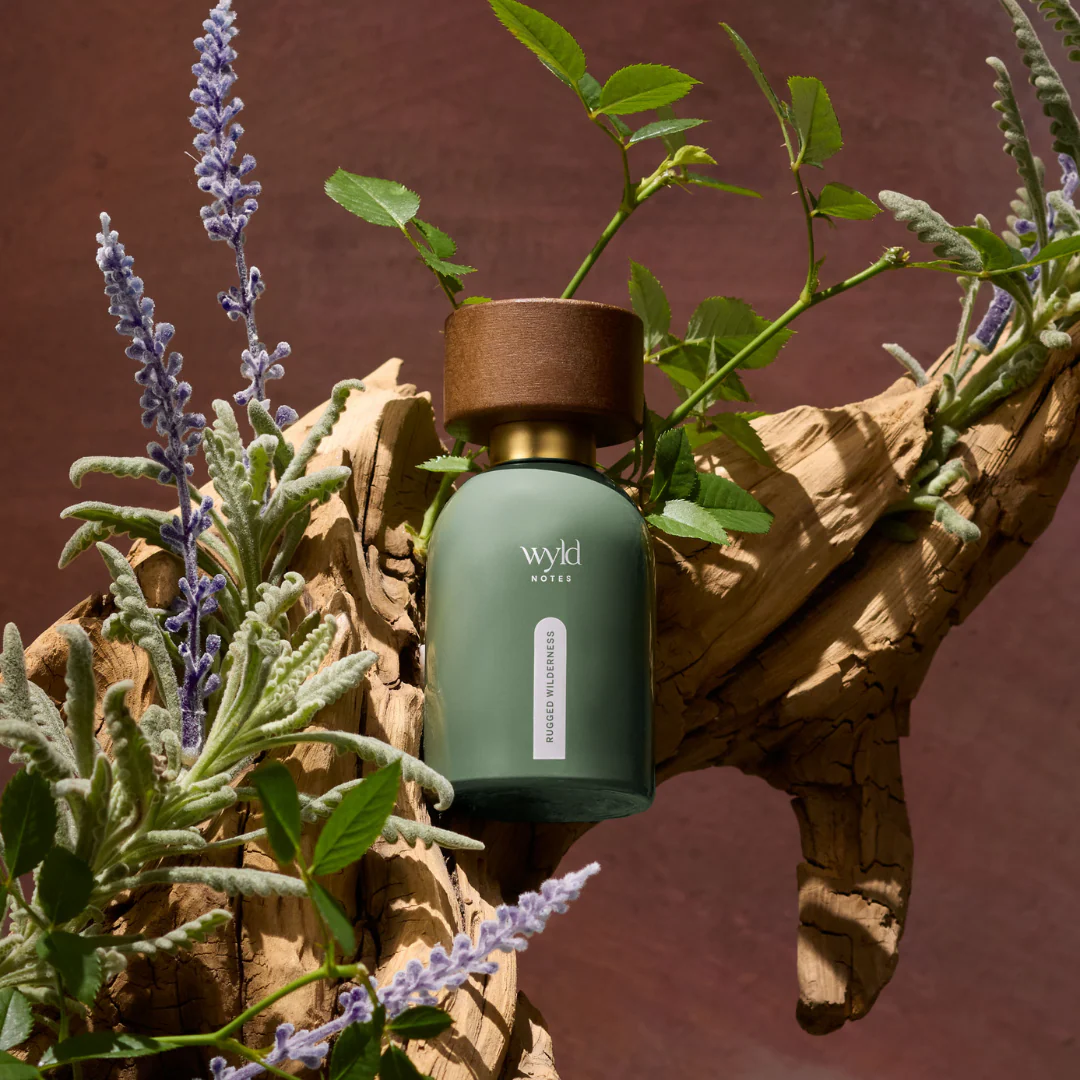 Green bottle labeled "wyld notes rugged wilderness" on driftwood with lavender and leaves against a maroon background, evoking a natural, earthy mood.