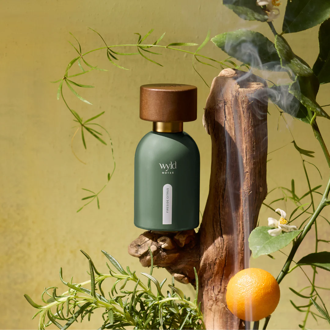 Green bottle labeled "wyld notes: fireside citrus" on wood with smoke, greenery, and an orange in a sunlit, earthy-toned setting.