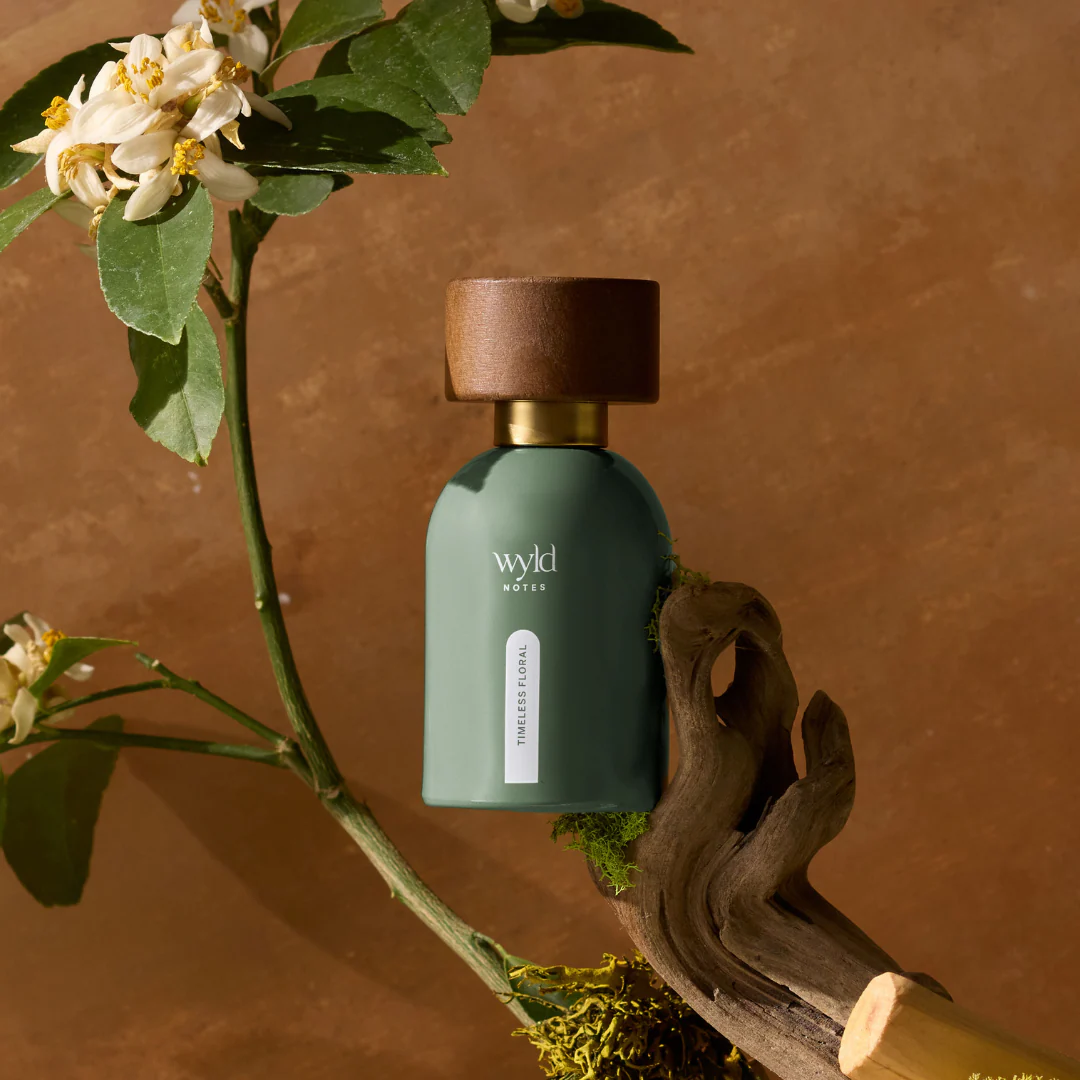 Perfume bottle labeled "Wyld Notes Timeless Floral" with a wooden cap, set against a brown background. Green branches and white flowers surround it.