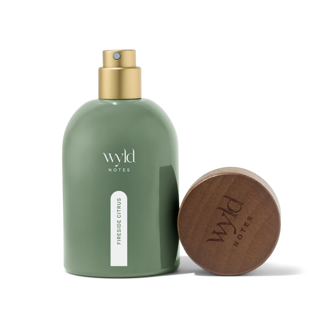 Green bottle with gold spray top, labeled "wyld notes fireside citrus." Wooden cap with matching logo. Text reads "Fireside Citrus." Minimalist design.