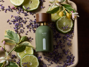 Green perfume bottle with "wyld NOTES" and "RUGGED WILDERNESS" text, surrounded by sliced limes, lavender petals, and lemon blossoms on a tray.