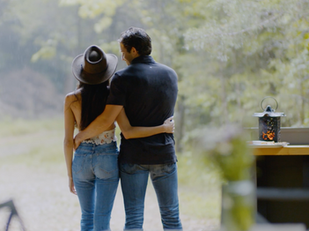 A couple embraces in a woodland setting, one wearing a wide-brim hat. There's a lantern on a counter. Peaceful, rustic ambiance.