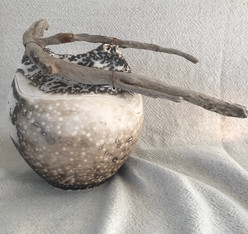 Cette urne funéraire de style obvara est en céramique. Elle doit son caractère organique à ses formes, aux couleurs de terre et à la branche de bois flotté qui l'orne au sommet.