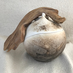 Cette urne funéraire doit son caractère unique au bois flotté qui la surmonte et l'enveloppe. Elle est en céramique de style obvara. La couleur brun beige et les formes sont organiques.