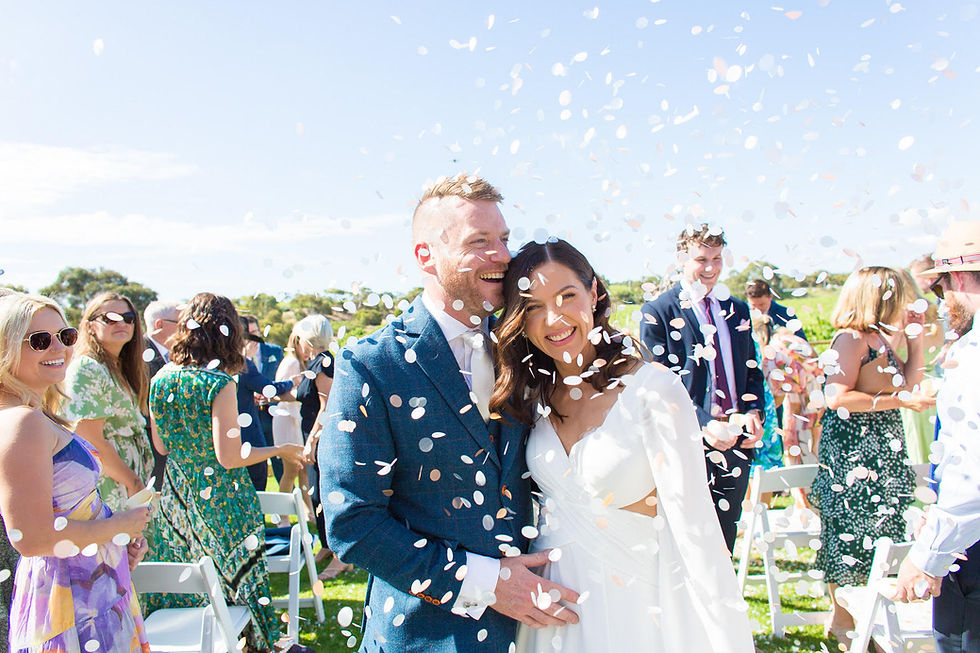 Smiling couple in wedding attire surrounded by confetti. Guests celebrate outdoors under clear blue sky. Joyful, festive atmosphere.