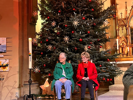 Zwei Mitarbeiterinnen sitzen auf zwei Stühlen vor einem dunkelgrünen, festlich geschmücktem Weihnachtsbaum in einer Kirche.