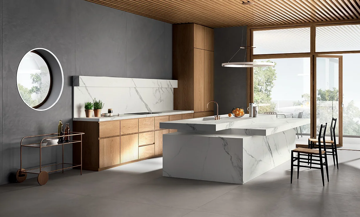 A luxurious kitchen island made entirely of Calacatta Venato porcelain slab, fully wrapped in the stone for a seamless, sculptural look. The slab features soft gray veining on a crisp white background, creating an elegant and timeless aesthetic. The monolithic design of the island serves as a stunning centerpiece, complementing the modern kitchen’s sleek cabinetry and refined finishes.