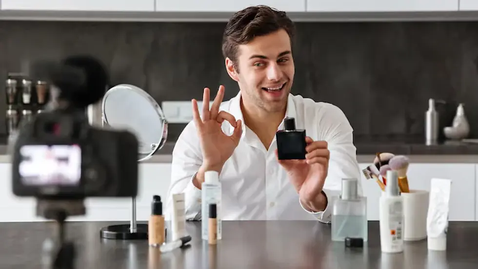Man in white shirt holding a perfume bottle, gesturing OK, smiling at camera. Makeup products on table, modern kitchen background. Mood is positive.
