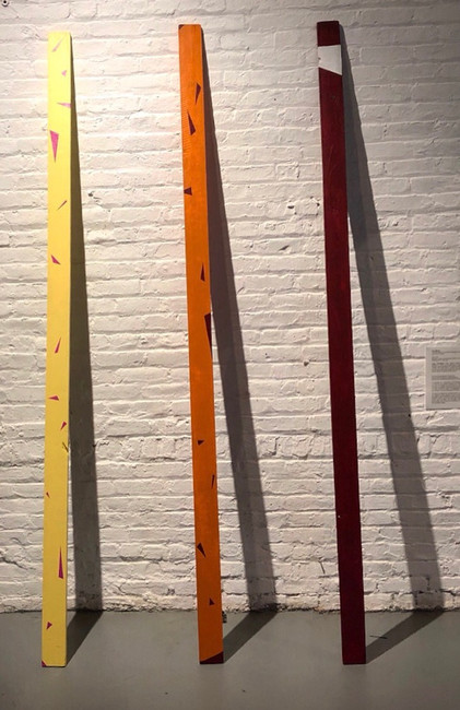Three painted wooden planks leaning and centered on a wall. The left most plank is light yellow with magenta triangles painted throughout the surface seemingly floating, falling, or crashing. The center plank is orange with deep red triangles on the surface seemingly floating, falling, and crashing. The right most plank is deep red with a white band near the top. Shadows casted from each plank make three large triangular structures
