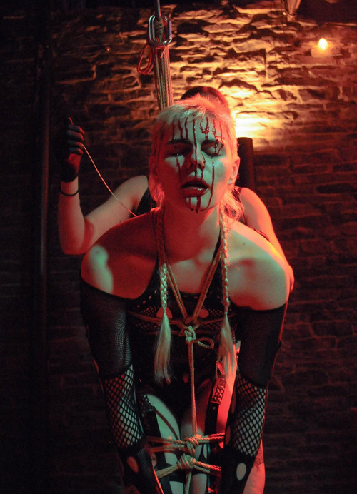 A white person is bent over, tied from their chest to their thighs while bleeding from their forehead. The blood drips down their face all the way to their chin. Behind them is another white person with one black latex glove in the air holding some rope. 