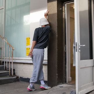 Jessica Karuhanga is leaning on a wall between an outdoor enclosed stairway and an exterior door. Her body is twisted against the door frame, with her face hidden and her hands open, her right arm up, her left hand down, and her legs crossed at the knee. Jessica is a slender, brown-skinned, femme-identified woman wearing a fuzzy white Kangol-style hat, white corded earbuds, a large black Tracy Chapman shirt tucked into loose light-colored sweatpants, gathered at the ankle with white socks and colorful sneakers.