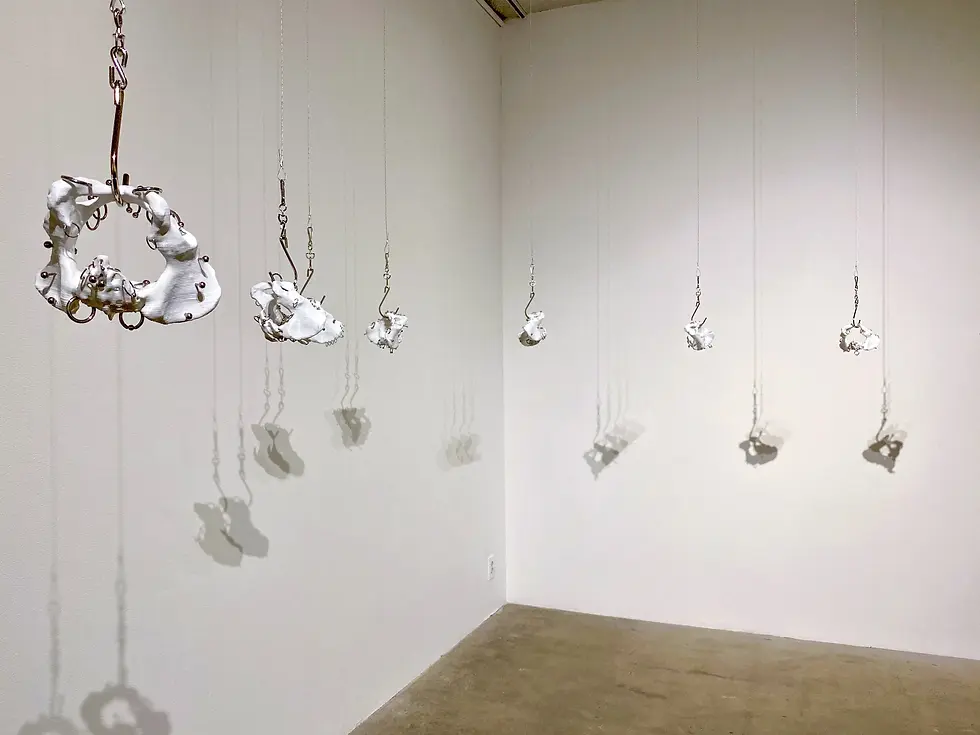 New Artifacts (2020) consists of eleven sculptural works made from filament, metal hardware, and sterling silver hardware. In this installation view, several irregular white forms resembling fragmented or distorted anatomical structures hang from the ceiling at varying heights on thin wires and metal hooks. The forms range in size and are pierced, encircled, and connected by small rings, bars, and other pieces of hardware. Directional lighting casts elongated shadows of the suspended sculptures across two white gallery walls.