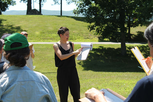 A white performer gestures towards the open diagram on their clipboard to a group of participants in a sunny pasture.