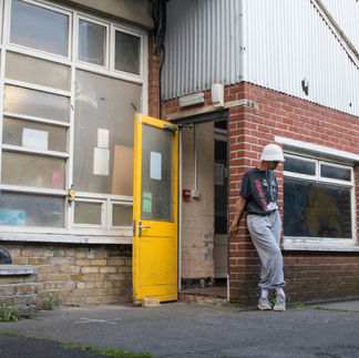Jessica Karuhanga is near a yellow exterior door, leaning against a brick wall, holding the corner of the wall while facing away from the corner, with her legs crossed at the knee. Jessica is a slender, brown-skinned, femme-identified woman wearing a fuzzy white Kangol-style hat, white corded earbuds, a large black Tracy Chapman shirt tucked into loose light-colored sweatpants, gathered at the ankle with white socks and colorful sneakers.