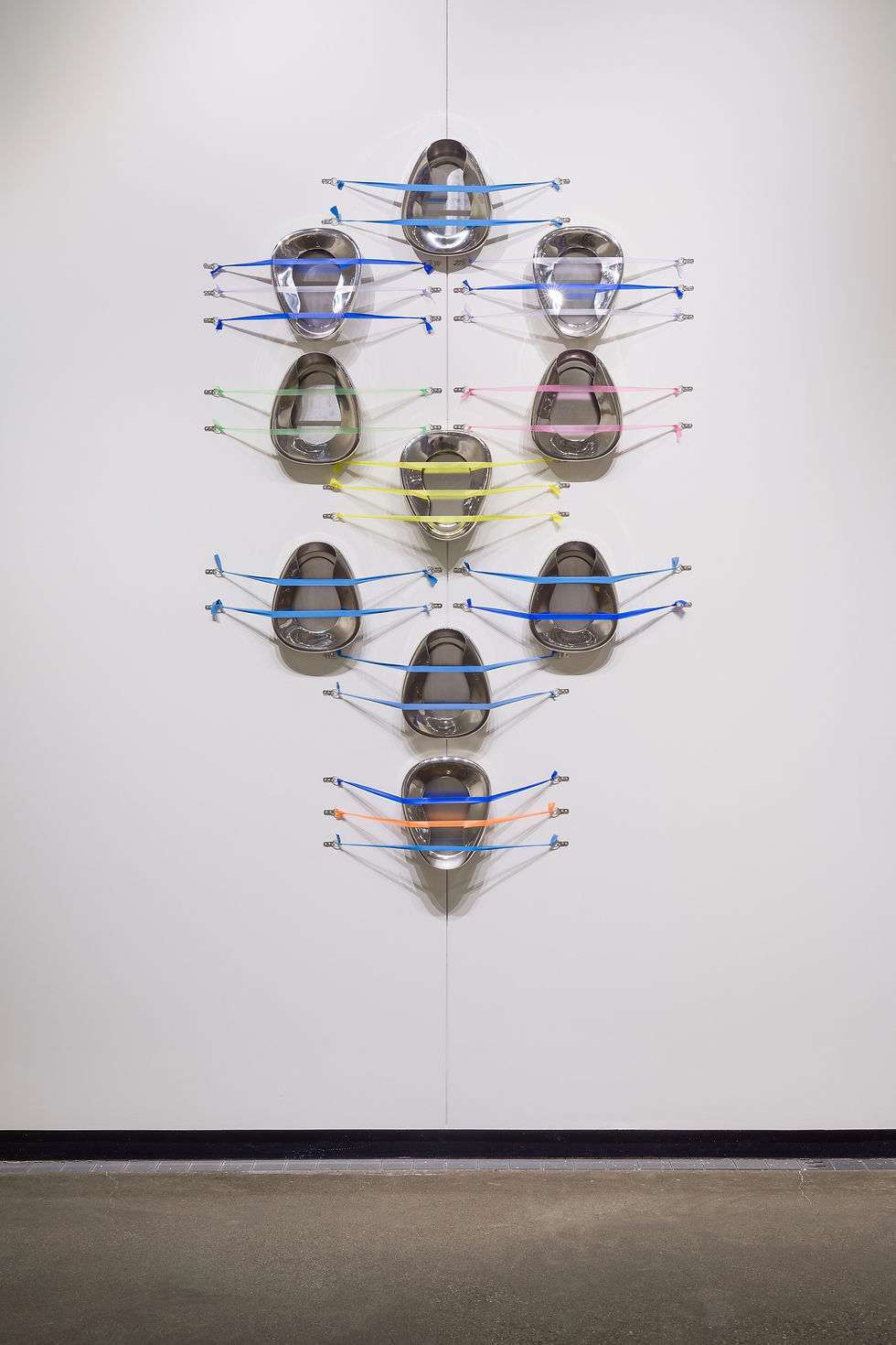 Twelve silver bedpans, oriented vertically, are tethered to the wall by taut colored tourniquets stretched laterally across them, two or three per bedpan. The tourniquets are blue, yellow, orange, and green. The bedpans are arranged in the pattern of the Jewish Sefirot, or Tree of Life, with one at the top flanked by two slightly below, two bedpans are directly below these flanking ones, with a centered one slightle below these. Another set of three below this last group echoes the shape, with the central bedpan lower down, and there is one centered bedpan at the base of the pattern.