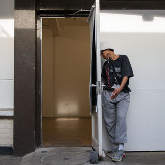 Jessica Karuhanga is leaning into and resting her head on an exterior door leading into an empty space with a window casting light onto the wall and floor. Jessica is a slender, brown-skinned, femme-identified woman wearing a fuzzy white Kangol-style hat, white corded earbuds, a large black Tracy Chapman shirt tucked into loose light-colored sweatpants, gathered at the ankle with white socks and colorful sneakers.