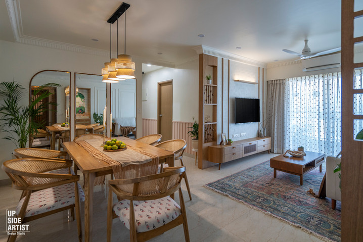 Spacious open-plan apartment interior displaying a wooden dining table under tiered pendant lights and a warm living room setup with a TV wall and traditional patterned rug.