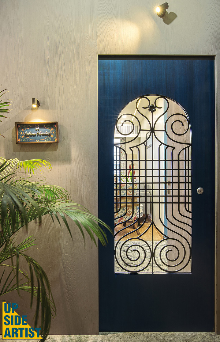 Front entrance of a home showing a deep blue wooden door with an arched cutout protected by decorative black iron scrollwork, positioned next to a nameplate reading 'Archana & Sandeep'.