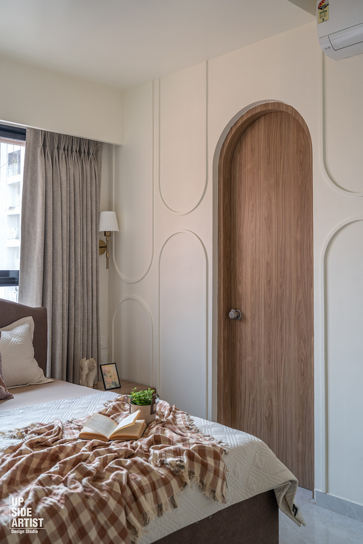 Bedroom corner view showing a walnut wood door under a custom drywall arch, accented by white wall moldings with elongated oval frames, a neutral bed, and brown plaid blanket.