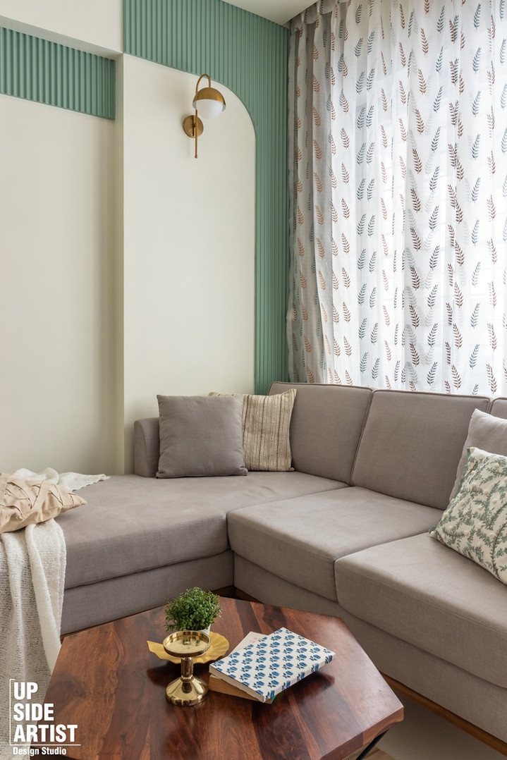 A living room seating arrangement featuring a comfortable grey fabric sectional sofa styled with throw pillows, a custom walnut wood coffee table, a white tasseled blanket throw, and sheer white curtains with a black branch pattern print.