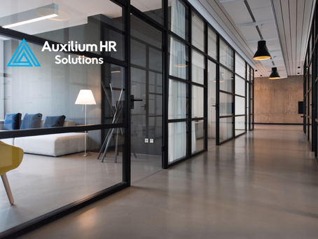 Modern office hallway with glass-walled meeting rooms, minimalist furniture, and the Auxilium HR Solutions logo in the foreground.
