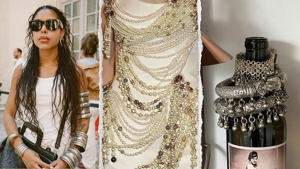 Stylish woman in sunglasses, arm cuffs, white top holding a black bag. Pearl and jewel-decorated fabric. Silver jewelry on a bottle in the background.