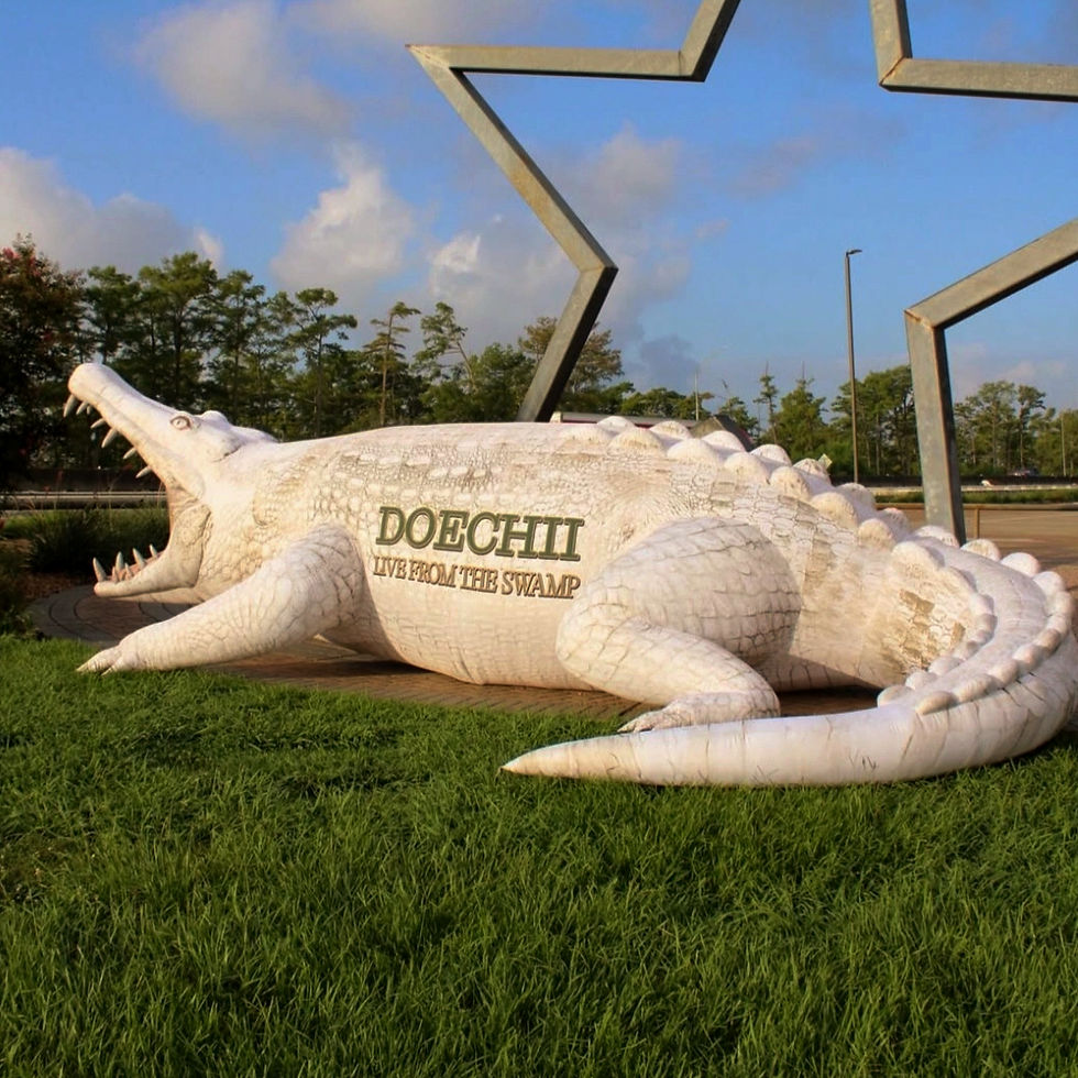 Large white crocodile balloon on grass with text "DOECHII LIVE FROM THE SWAMP." Metal star shape and trees in sunny background.