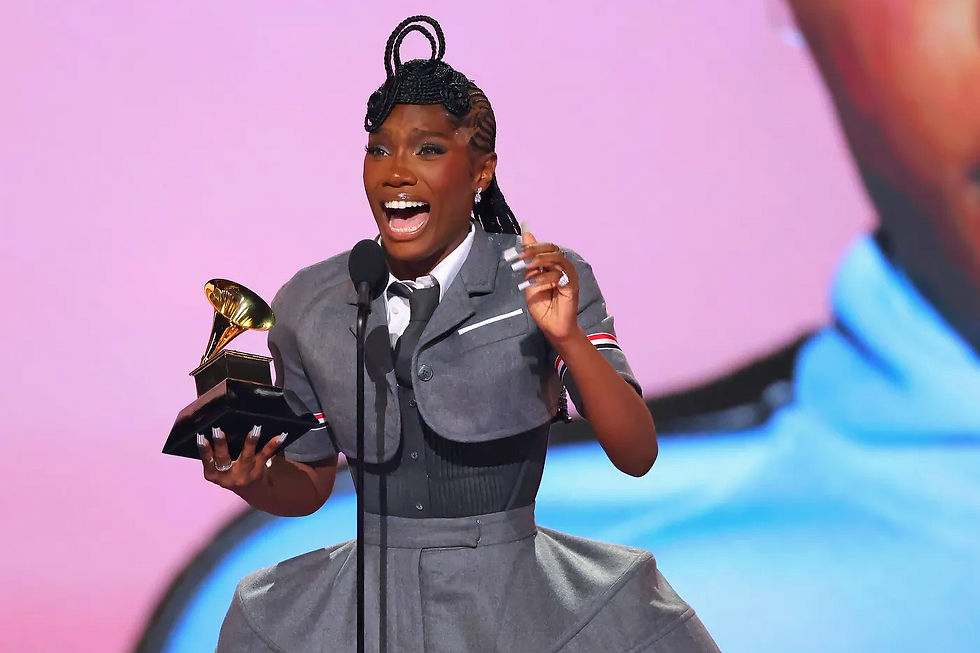 A person in a gray outfit smiles widely, holding a Grammy award on stage with a pink background. They appear joyful and excited.