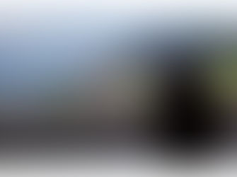 Person in striped shirt films a village with green fields and mountains in the background. Overcast sky.  